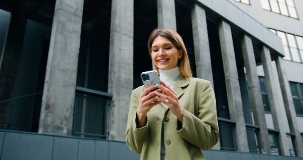 Successful young business woman uses smartphone in business district. Caucasian female employee entrepreneur uses a smartphone on urban background - Photo, Image