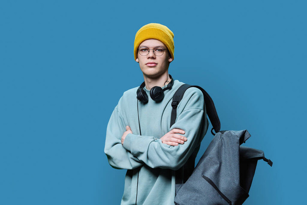 Young trendy hipster male in yellow knitted cap sweatshirt with headphones backpack looking at camera with crossed arms on blue studio background. Youth, fashion, leisure, lifestyle concept - Foto, afbeelding