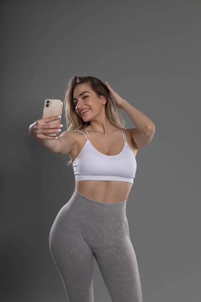 junge attraktive Fitness-Mädchen posiert und macht Selfie im Studio mit grauem Hintergrund - Foto, Bild