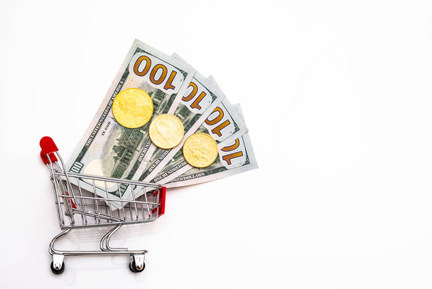 carrinho de compras sobre rodas com moedas de ouro e dólares de papel em um fundo branco - Foto, Imagem