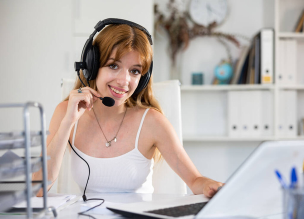 Attractive young woman using headphones with microphone and laptop while working in call center. - Foto, afbeelding