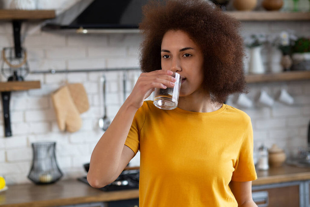 Šťastná africká Američanka se sklenicí čerstvé vody v bílém kuchyňském portrétu. Krásná usmívající se africká americká dívka pití čisté minerální vody - Fotografie, Obrázek