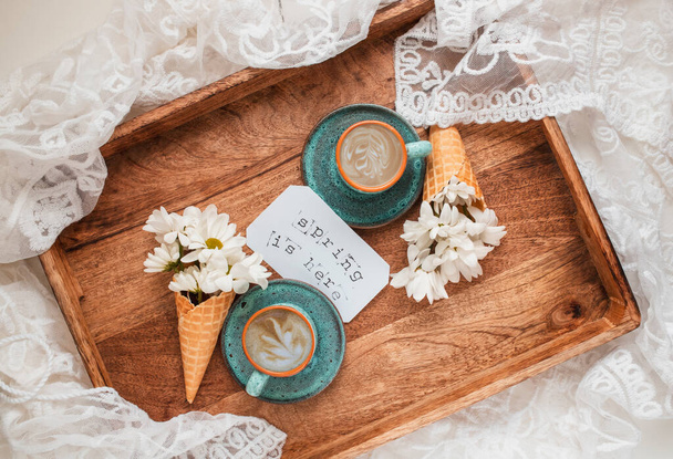 Romantic flat lay composition with two cup of coffee, white flowers in a waffle cones, label with text "Spring is here" on wooden tray. Good morning with a coffee in bed.  - Foto, immagini