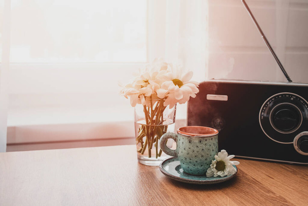 Eine Tasse Kaffee, ein Radio und ein Glas mit Frühlingsblumen auf dem Holztisch neben dem Fenster. Frühlingshafte Stimmung und gemütliches Wohnkonzept.  - Foto, Bild