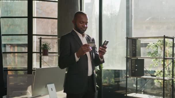 African American ώριμος άνθρωπος στο ηλιόλουστο γραφείο κατέχουν κινητό τηλέφωνο αρσενικό επιχειρηματία που πληρώνει online παραγγελία online ψώνια e-banking app στείλετε χρήματα πληρώσει χρήση τραπεζική πιστωτική κάρτα e-commerce οικονομική εύκολη πληρωμή - Πλάνα, βίντεο