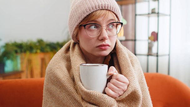 Kranke junge Frau mit Hut in Plaid gehüllt sitzt allein zitternd vor Kälte auf dem Sofa und trinkt heißen Tee in einer unbeheizten Wohnung. Ungesundes krankes Mädchen in Brille fühlt sich unwohl und versucht, sich zu Hause aufzuwärmen - Foto, Bild