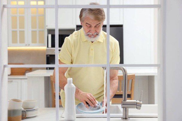 Üst düzey adam mutfak lavabosunun üstündeki tabağı yıkıyor. Dışarıdan görünüyor. - Fotoğraf, Görsel