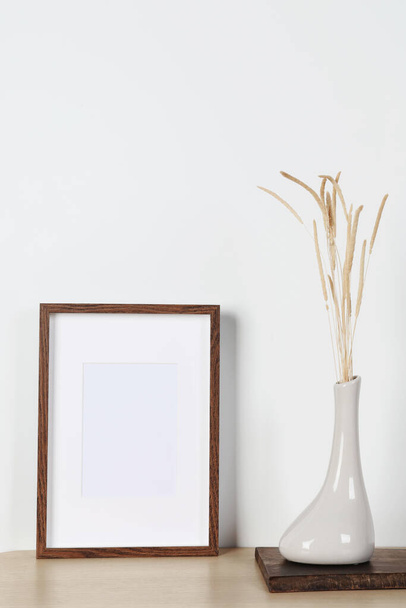 Empty photo frame and vase with dry decorative spikes on wooden table. Mockup for design - Fotó, kép