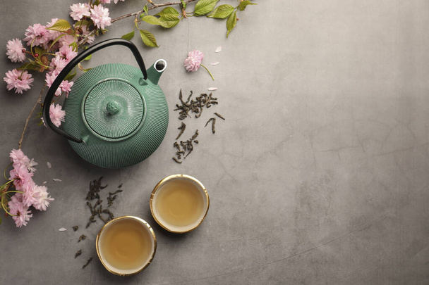 Traditional ceremony. Cups of brewed tea, teapot and sakura flowers on grey table, flat lay with space for text - Foto, afbeelding