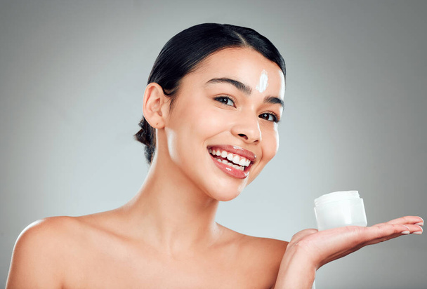 Studio Portrait of a beautiful smiling mixed race woman applying cream to her face. Hispanic model with glowing skin holding moisturiser against a grey copyspace background. - Zdjęcie, obraz