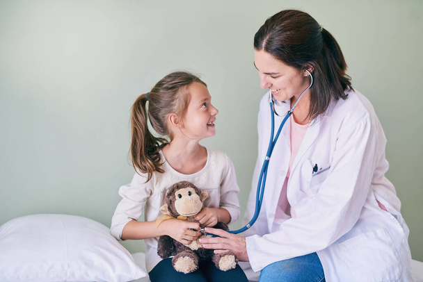 Medical, kids and a girl at a woman pediatrician for an appointment or checkup in the hospital. Healthcare, stethoscope and teddy bear with an adorable female child sitting on a bed in the clinic. - 写真・画像