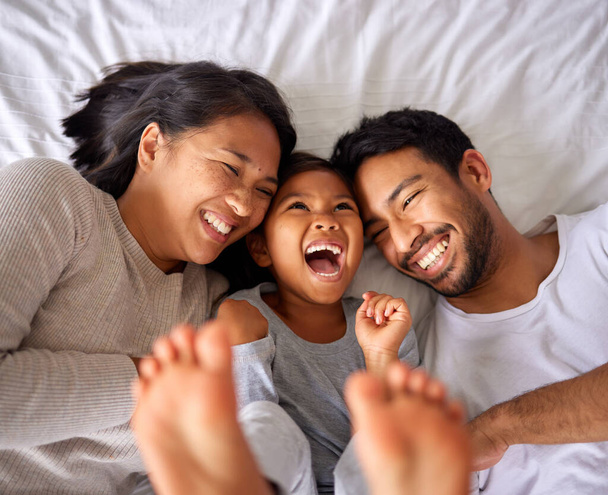 Ethnic family resting in bed together. Young happy couple laughing while tickling their happy little girl in a big soft bed at home - Fotoğraf, Görsel