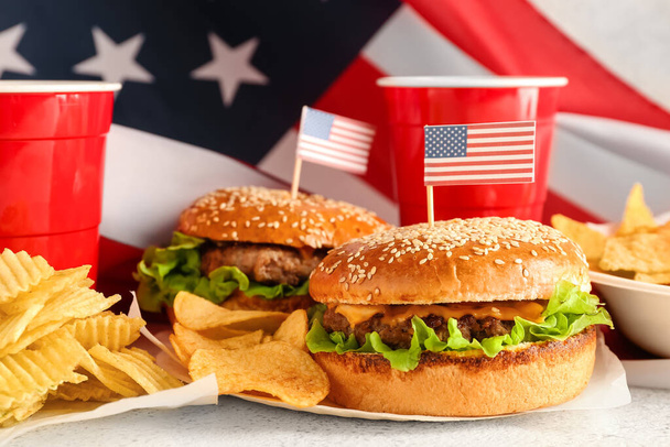 Tasty burgers, potato chips, cups of drink and American flag on light background, closeup. Memorial Day celebration - Foto, Imagem