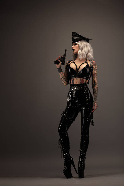 Pretty tattooed young woman wearing latex lingerie, cap and gun - Photo, Image