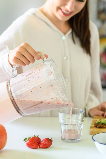 Crop positive young female in casual white shirt pouring fresh strawberry smoothie into transparent glass and smiling while preparing healthy breakfast in kitchen - Valokuva, kuva