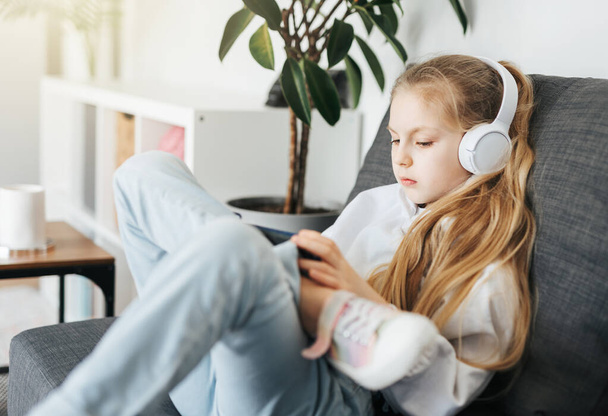 Little girl  lying on the couch with an electronic tablet listens to music in white headphones. Using wireless devices for education. - Valokuva, kuva