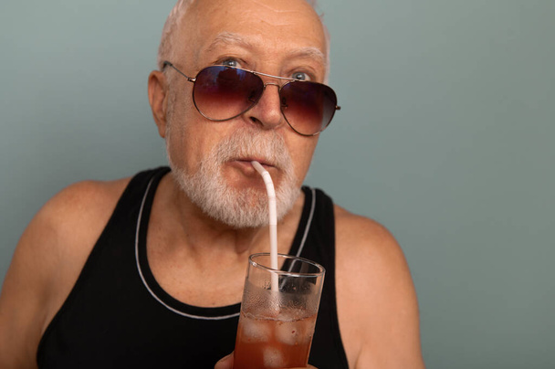 Ein charismatischer älterer Mann mit Sonnenbrille und schwarzem T-Shirt hält ein eisiges Getränk in der Hand, einen interessanten Gesichtsausdruck, einen hellen Hintergrund, einen Platz für Text. - Foto, Bild
