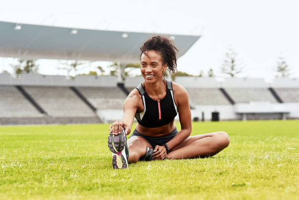Happy woman, athlete and stretching legs on grass at stadium getting ready for running, workout or exercise. Active female person or runner in warm up leg stretch, athletic fitness or sports on field. - Zdjęcie, obraz