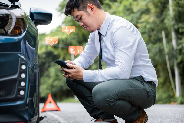 Businessman calling roadside assistance after his car broke down on the road. Waiting for help and support, he sits by his car wearing a white shirt. Concept of roadside assistance and car accident - 写真・画像