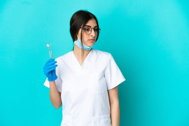 Молода біла жінка-стоматолог тримає інструменти ізольовані на синьому фоні, дивлячись в сторону
 - Фото, зображення