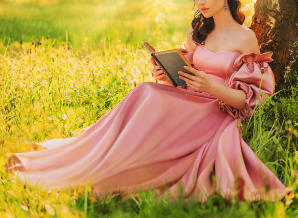 Fantasy woman sits under tree holding romantic book in hands reading novel smiling face, lips close-up. Pink long dress. Fairy girl in summer nature green grass magic sun light. art photo real people - Photo, image
