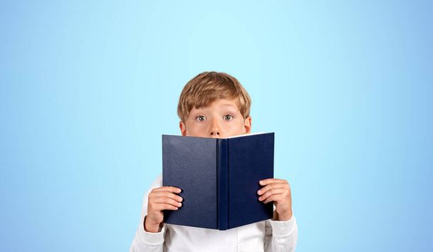 Portrait of surprised little boy covering his face with book standing over blue background. Concept of education, reading and gaining knowledge for future - Photo, Image