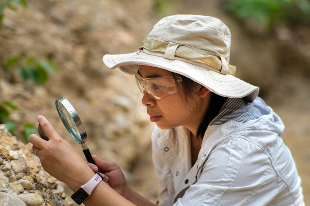 Female geologist using magnifying glass to examine and analyze rock, soil, sand in nature. Archaeologists explore the field. Environmental and ecology research. - Photo, Image