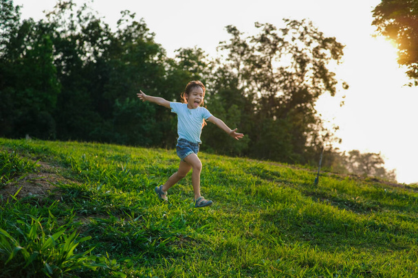 Happy little girl running on the grass in the summer park. Little girl running through the meadow with sunset light. - Photo, Image