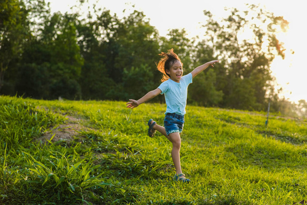 Happy little girl running on the grass in the summer park. Little girl running through the meadow with sunset light. - Фото, изображение