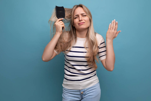 μελαγχολική ξανθιά γυναίκα με δυσκολία χτενίζει τα μαλλιά της σε μπλε φόντο. - Φωτογραφία, εικόνα