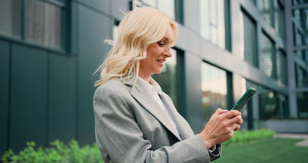 Portrait of happy blond business woman typing by mobile phone outdoors. Woman looking into smartphone screen chatting online or message or social media. - Fotografie, Obrázek