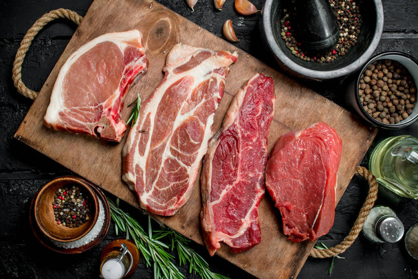 Raw meat. Pork and beef steaks on a tray with spices and rosemary sprig. On a black rustic background. - Photo, image