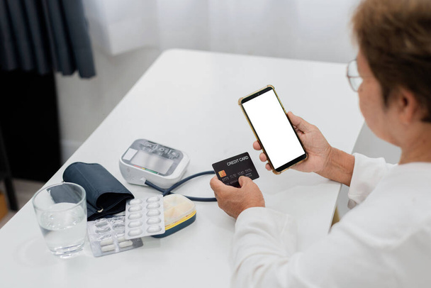 telemedicine έννοια, μια ηλικιωμένη Ασιάτισσα κρατά μια πιστωτική κάρτα και ένα smartphone, για να πληρώσει για ιατρικές συμβουλές, λευκή οθόνη και το μονοπάτι ψαλίδισμα. - Φωτογραφία, εικόνα