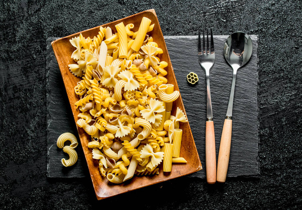Different pasta dry on a plate with fork and spoon. On black rustic background - Foto, Imagem