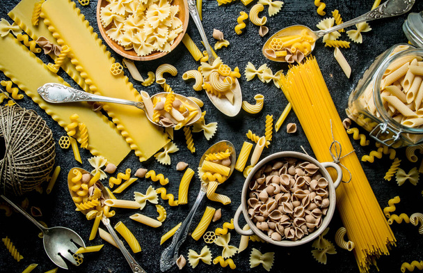 Pasta background. Assortment of different types of dry pasta in bowl and spoons with twine. On black rustic background - Foto, imagen