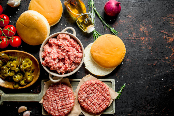 Raw burgers with ground beef, oil, buns and tomatoes. On dark rustic background - 写真・画像