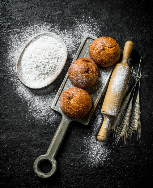 Fragrant buns with flour and rolling pin. On black rustic background - Photo, Image