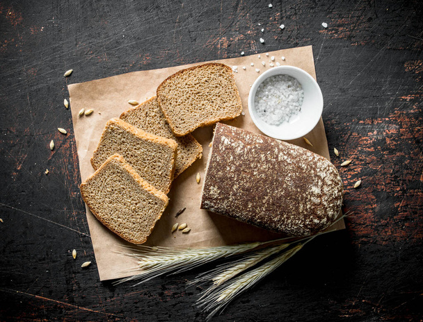 Gesneden brood op papier met zout en spikeletten. Op donkere rustieke achtergrond - Foto, afbeelding
