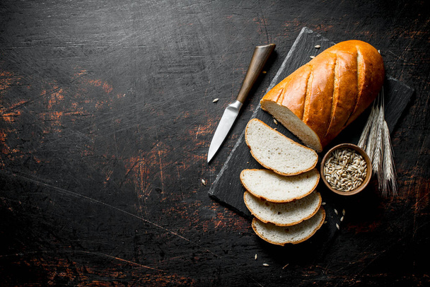Pieces of bread with a knife, grain and spikelets. On dark rustic background - Zdjęcie, obraz