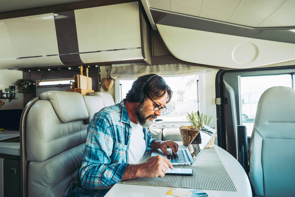  man using computer and phone sitting indoor inside camper van modern vehicle. People enjoying technology and connection in travel vacation lifestyle. Living vanlife and working. New job office - Fotografie, Obrázek