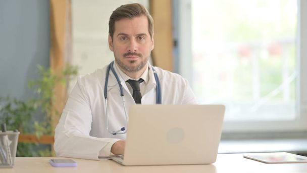Médecin masculin travaillant sur ordinateur portable regardant vers la caméra - Photo, image