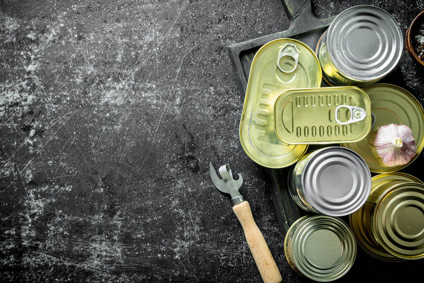 Set of different kinds of tin cans with canned food on a cutting Board with garlic and a opener. On dark rustic background - 写真・画像