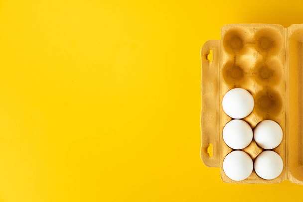 Ploché snášky bílých vajec v papírové hnědé krabici na žlutém pozadí. Horní pohled. Snídaně v ranní koncepci s kopírovacím prostorem pro volný text. - Fotografie, Obrázek
