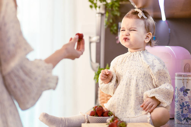 Mom feeding her baby girl with sour strawberries in the kitchen. Making sour face - 写真・画像