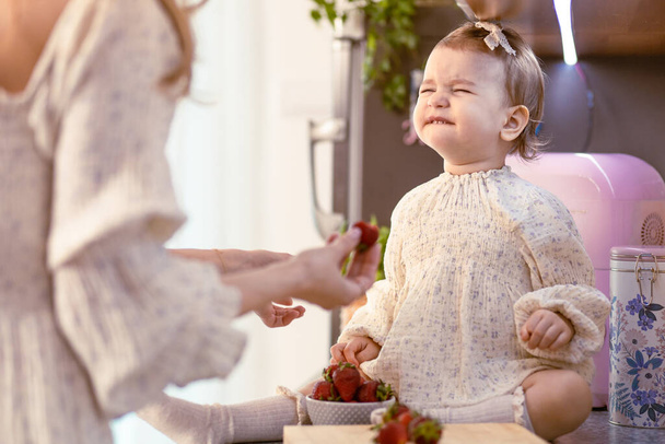 Mom feeding her baby girl with sour strawberries in the kitchen. Making sour face - Fotografie, Obrázek