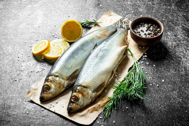 Salted herring on paper with lemon slices, dill and spices. On dark rustic background - Фото, изображение