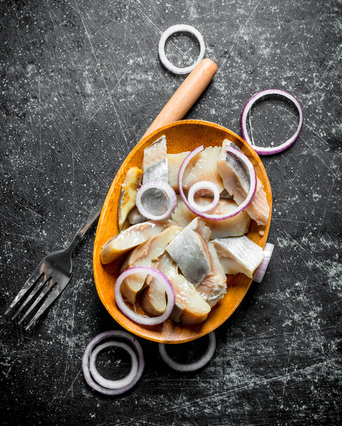 Pieces of salted herring on a plate with a fork and sliced onions. On dark rustic background - Фото, зображення