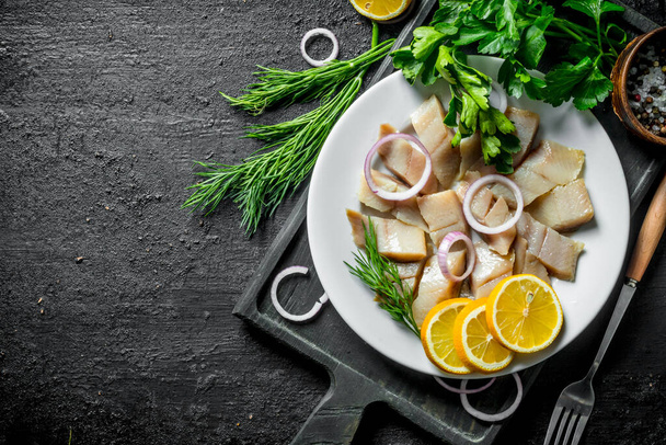 Pieces of salted herring with herbs, spices and lemon. On black rustic background - 写真・画像