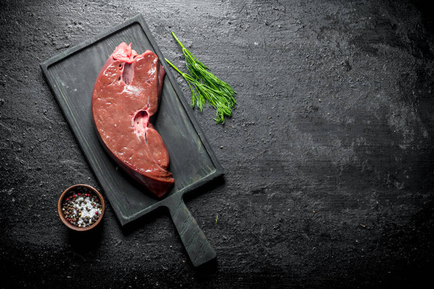 Piece of raw liver on a cutting Board with dill and spices. On black rustic background - Foto, immagini
