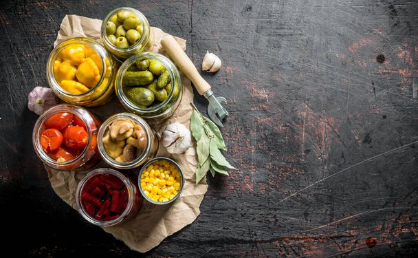 Pickled vegetables in jars on the paper with the Bay leaves and the cloves of garlic. On dark rustic background - Photo, image